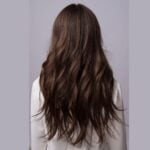 Tips on How to Style Hair Extensions to Look Natural A person with long, wavy brown hair-styled with natural hair extensions-is shown from the back, wearing a white top against a plain light grey background.