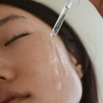 A close-up of a person applying facial serum with a dropper on their cheek, eyes closed, showing clear liquid on their skin.