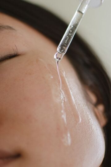 A close-up of a person applying facial serum with a dropper on their cheek, eyes closed, showing clear liquid on their skin.