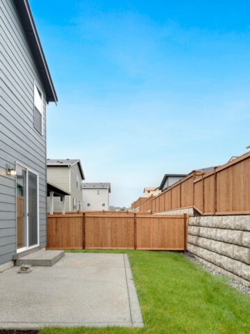 A small back garden with green grass, a concrete patio, a wooden fence installed by one of the top fencing companies Austin has to offer, and part of a grey two-storey house on the left under a clear blue sky.
