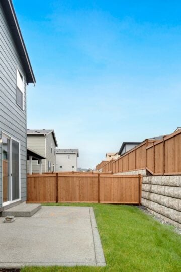 A small back garden with green grass, a concrete patio, a wooden fence installed by one of the top fencing companies Austin has to offer, and part of a grey two-storey house on the left under a clear blue sky.
