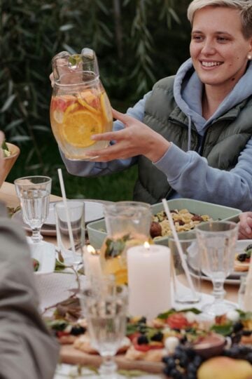 A person sits at a garden table with food, holding a jug of citrus water. Others raise glasses, and the table is set with candles, salads, and fruit—a perfect scene for outdoor entertaining.