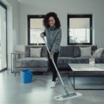 The Best House Cleaning Companies in Houston, TX A woman wearing headphones mops the floor in a modern living room, showcasing efficient house cleaning with a blue bucket nearby and a grey sofa in the background.