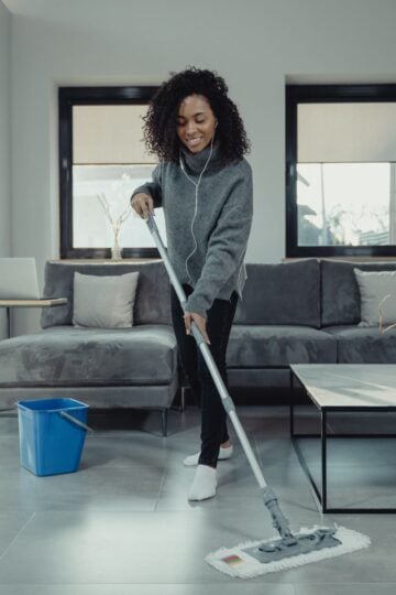 A woman wearing headphones mops the floor in a modern living room, showcasing efficient house cleaning with a blue bucket nearby and a grey sofa in the background.