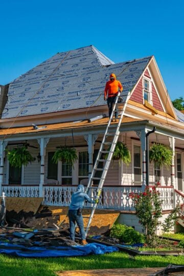 Two workers repair the roof of a house; one stands on a ladder while the other manages materials below. Roofing supplies and debris—common sights during an Austin roof replacement—are scattered across tarpaulins on the ground, showcasing the efficient teamwork typical of experienced residential roofing companies.