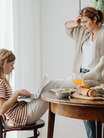 A woman stands by a table looking concerned-a scene that many mums may recognise from their parenting journey-while a girl types on a laptop, seated with her legs crossed. The table is set with food items, drinks, and crockery, capturing the often overwhelming and sometimes "touched out" moments of family life.