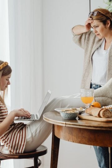 Why So Many Mums Reach the Point of Being Completely Touched Out A woman stands by a table looking concerned—a scene that many mums may recognise from their parenting journey—while a girl types on a laptop, seated with her legs crossed. The table is set with food items, drinks, and crockery, capturing the often overwhelming and sometimes "touched out" moments of family life.