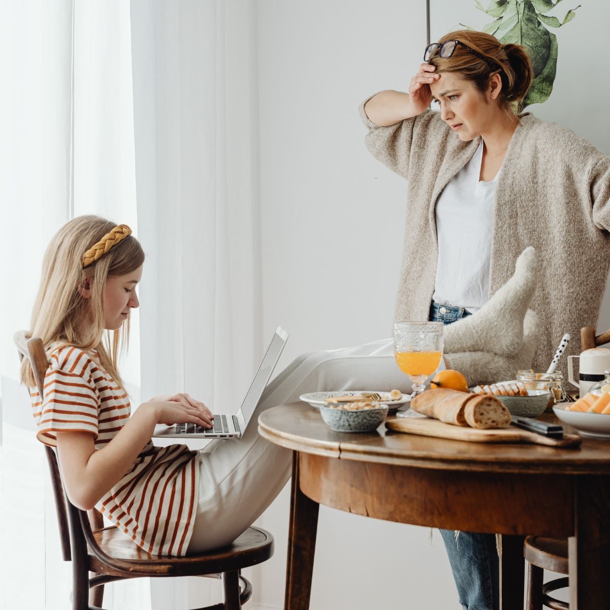 Why So Many Mums Reach the Point of Being Completely Touched Out A woman stands by a table looking concerned—a scene that many mums may recognise from their parenting journey—while a girl types on a laptop, seated with her legs crossed. The table is set with food items, drinks, and crockery, capturing the often overwhelming and sometimes "touched out" moments of family life.