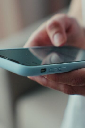 A person holds and uses a mobile phone with a light blue case, focusing on the screen and side buttons—perhaps preparing to teach your child about recognising a text scam in 2026.