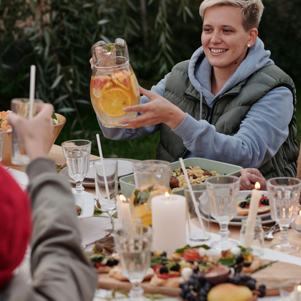 How to Create the Perfect Backyard for Entertaining Guests A person sits at a garden table with food, holding a jug of citrus water. Others raise glasses, and the table is set with candles, salads, and fruit—a perfect scene for outdoor entertaining.