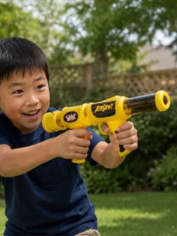 Smiling child with short dark hair holds a yellow and black Pop Blast foam ball blaster outside in a garden with grass, trees, and a wooden fence-children will love this playful adventure!.