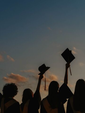 Four graduates in gowns hold up their mortarboards against a sunset sky, celebrating their achievement outdoors.