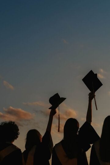 Four graduates in gowns hold up their mortarboards against a sunset sky, celebrating their achievement outdoors.