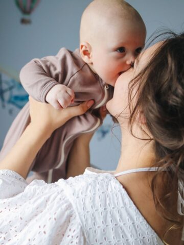Woman in a white blouse holds a baby in a brown babygrow, lifting the baby close to her face while they touch noses in a softly lit room, capturing a tender moment of emotional support and connection that every mother-especially during pregnancy-cherishes.