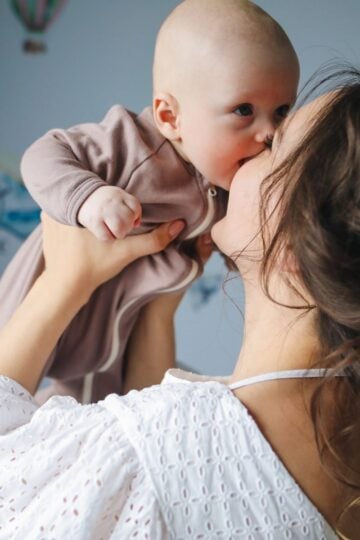 Woman in a white blouse holds a baby in a brown babygrow, lifting the baby close to her face while they touch noses in a softly lit room, capturing a tender moment of emotional support and connection that every mother—especially during pregnancy—cherishes.