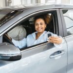 Woman sitting in the driver's seat of a car, smiling and holding up a car key-a moment of excitement many new drivers experience-while another person is partially visible in the passenger seat.