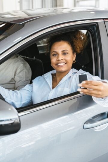 Woman sitting in the driver’s seat of a car, smiling and holding up a car key—a moment of excitement many new drivers experience—while another person is partially visible in the passenger seat.