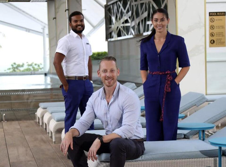 From Five-Star Luxury to Casual Retreats: How to Choose Hotel Uniforms That Reflect Your Brand Three people pose by a swimming pool; one man sits on a lounger while another man and a woman stand behind him, all dressed in business or professional attire.