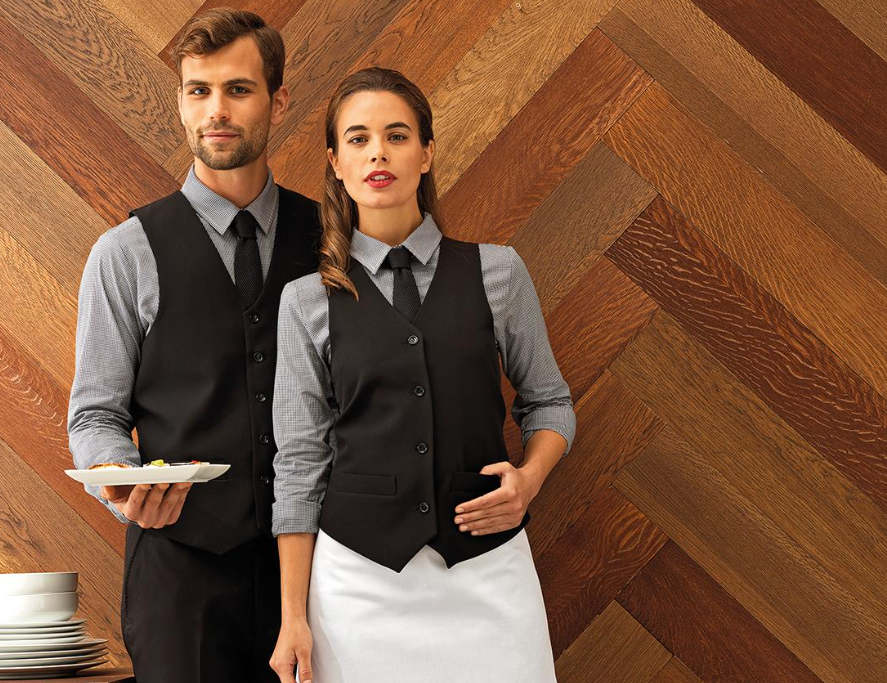 From Five-Star Luxury to Casual Retreats: How to Choose Hotel Uniforms That Reflect Your Brand Two restaurant waiters in matching uniforms stand against a wooden herringbone wall; one holds a plate of food, and the other wears a white apron.