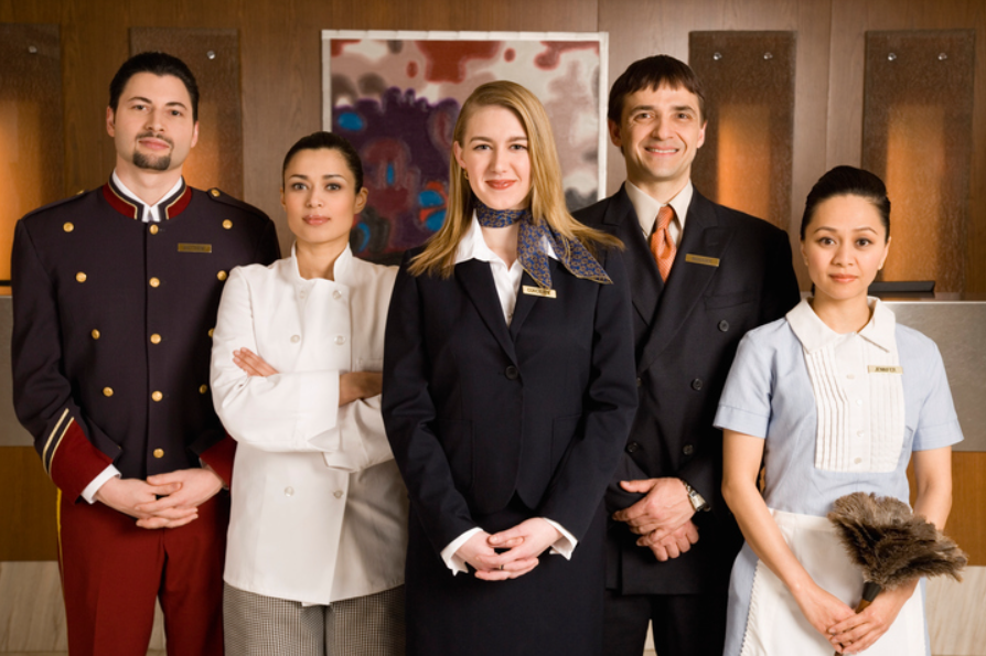 From Five-Star Luxury to Casual Retreats: How to Choose Hotel Uniforms That Reflect Your Brand Five hotel staff members in uniform stand side by side, facing the camera, against a modern wooden wall with artwork in the background.