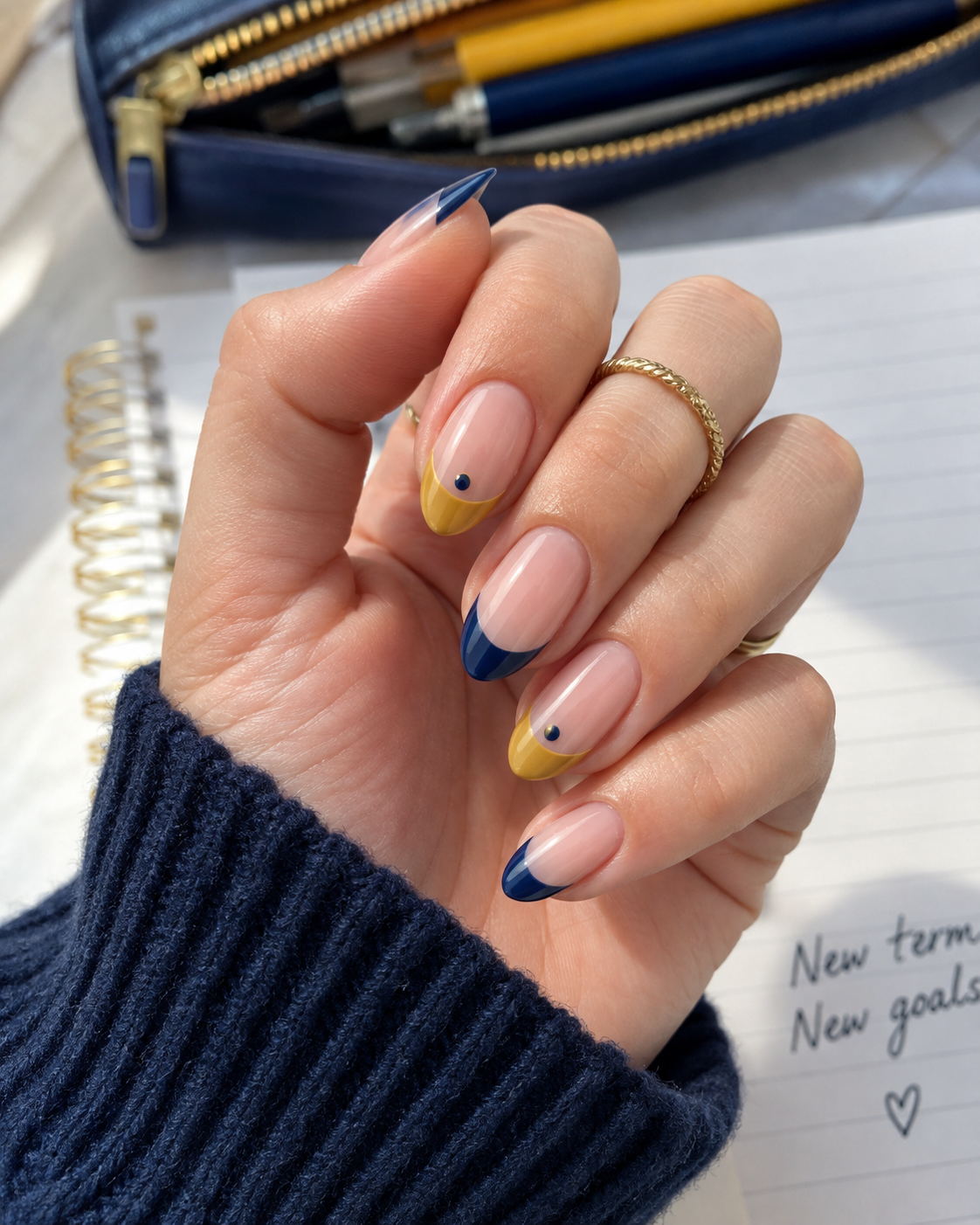 A hand with yellow and blue French tip nails, each featuring a single blue dot—perfect back to school nail designs—holds a notebook. The person wears gold rings and a blue jumper, with pens and a pencil case in the background.