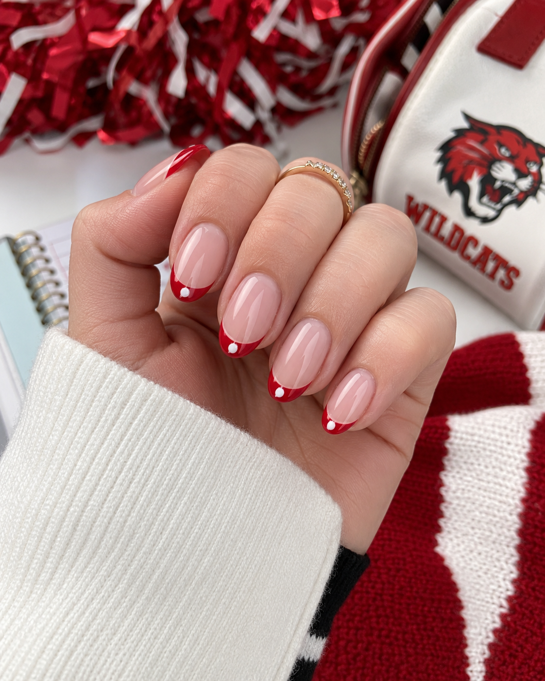 A hand with manicured nails featuring red French tips and white dots, showcasing one of the latest back to school nail designs, holds a spiral notebook; cheer pom-poms and a Wildcats bag are in the background.