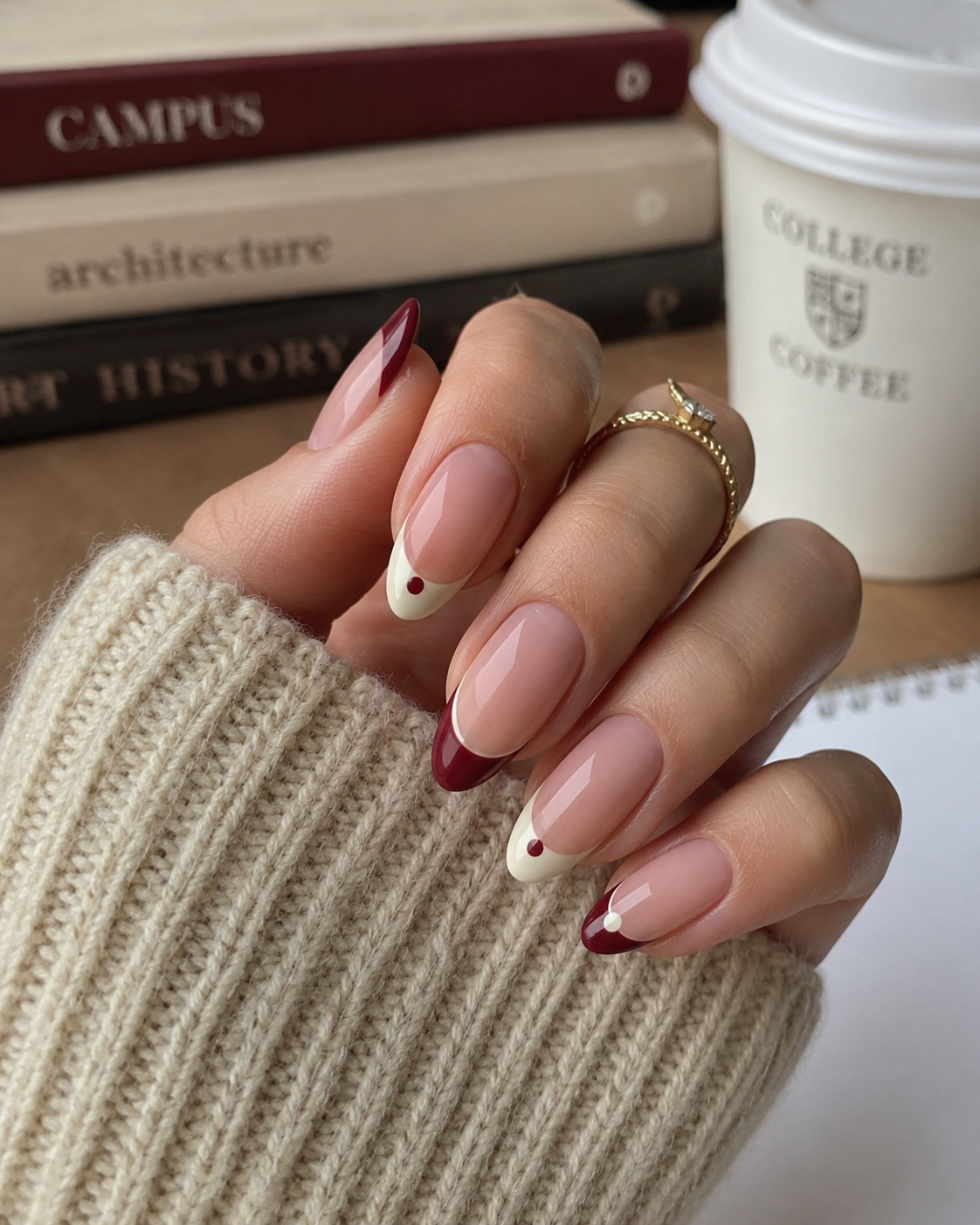 A hand with almond-shaped nails features red and beige French tips accented by red dots—perfect back to school nail designs. Books and a coffee cup are in the background, while the person wears a jumper and a gold ring.