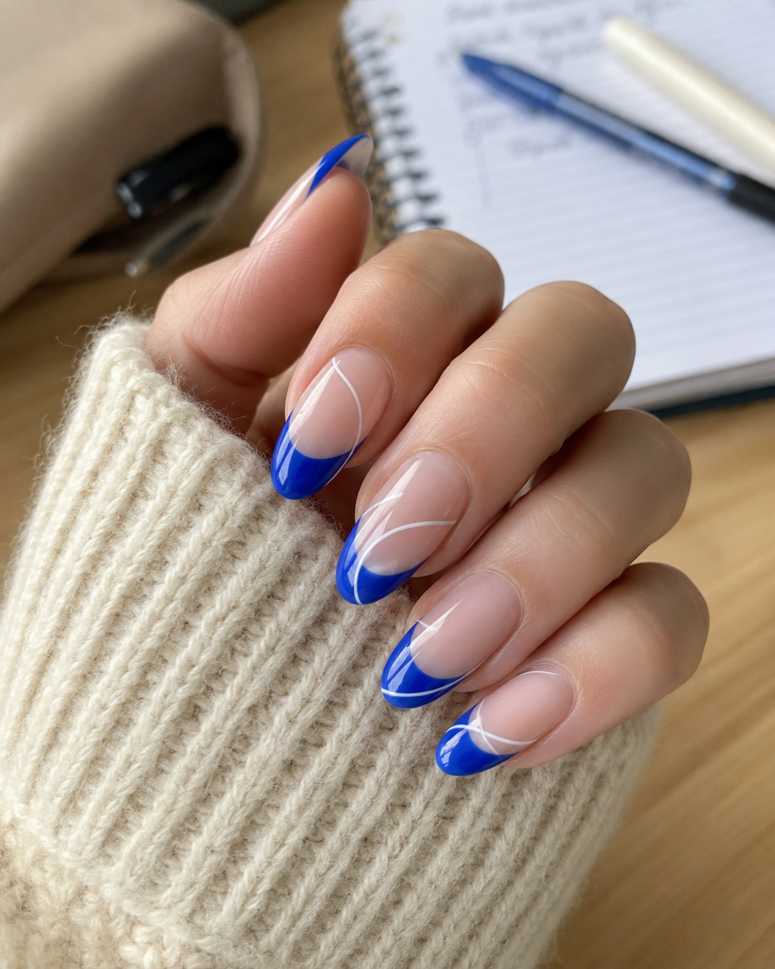 A hand with long nails featuring blue tips and white swirl designs rests on a beige jumper, with a pen and notebook in the background—perfect inspiration for stylish back to school nail designs.