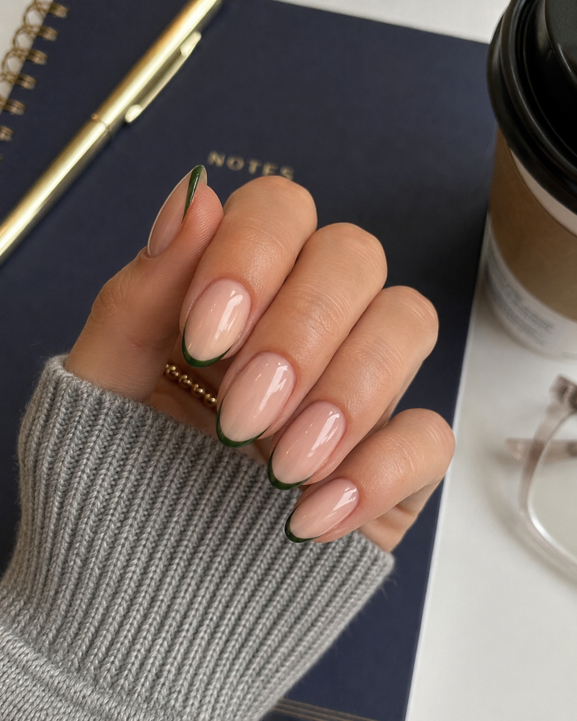 A hand with nude oval nails featuring thin green tips—an understated take on back to school nail designs—is resting on a navy notebook next to a coffee cup and spectacles.