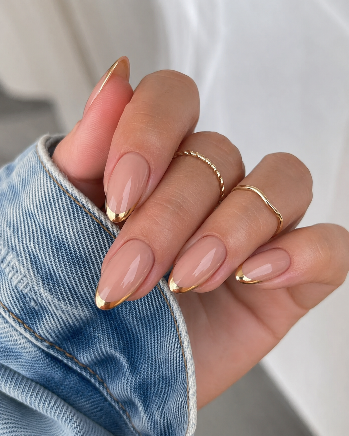 A hand with almond-shaped nails features a nude base and gold tips, perfect for chic back to school nail designs. The person wears two thin gold rings and a denim jacket.