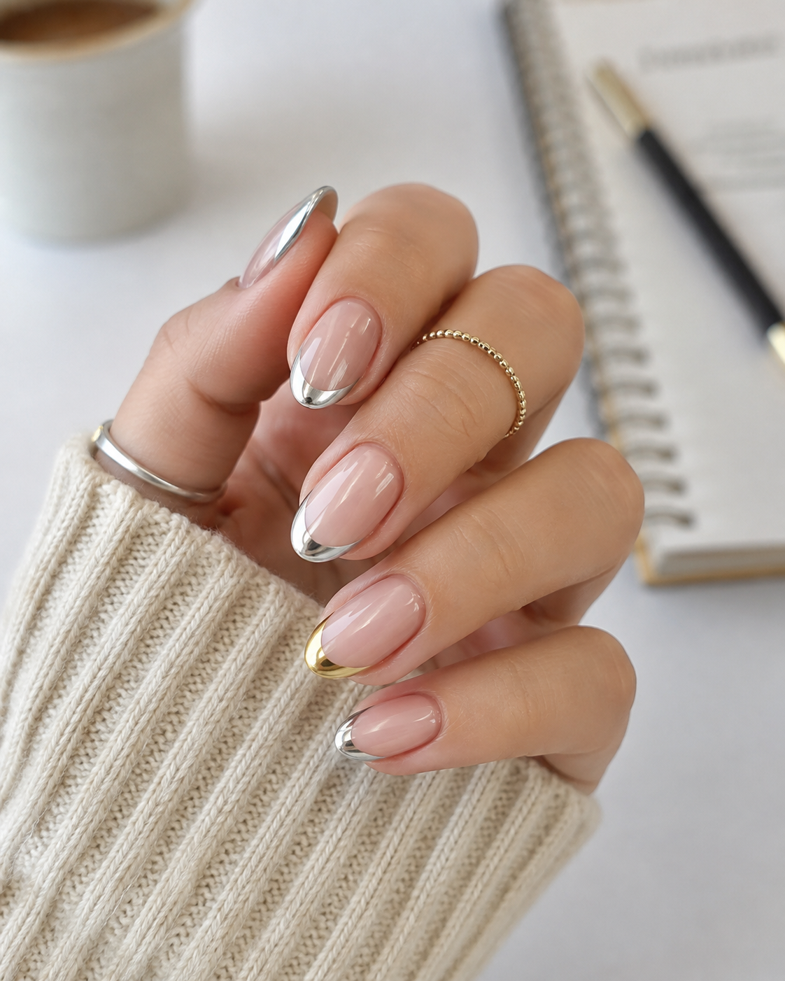 A hand with almond-shaped nails features a minimalist metallic French tip manicure, perfectly styled for back to school nail designs. The hand wears two rings and rests near a notebook, pen, and a cup.