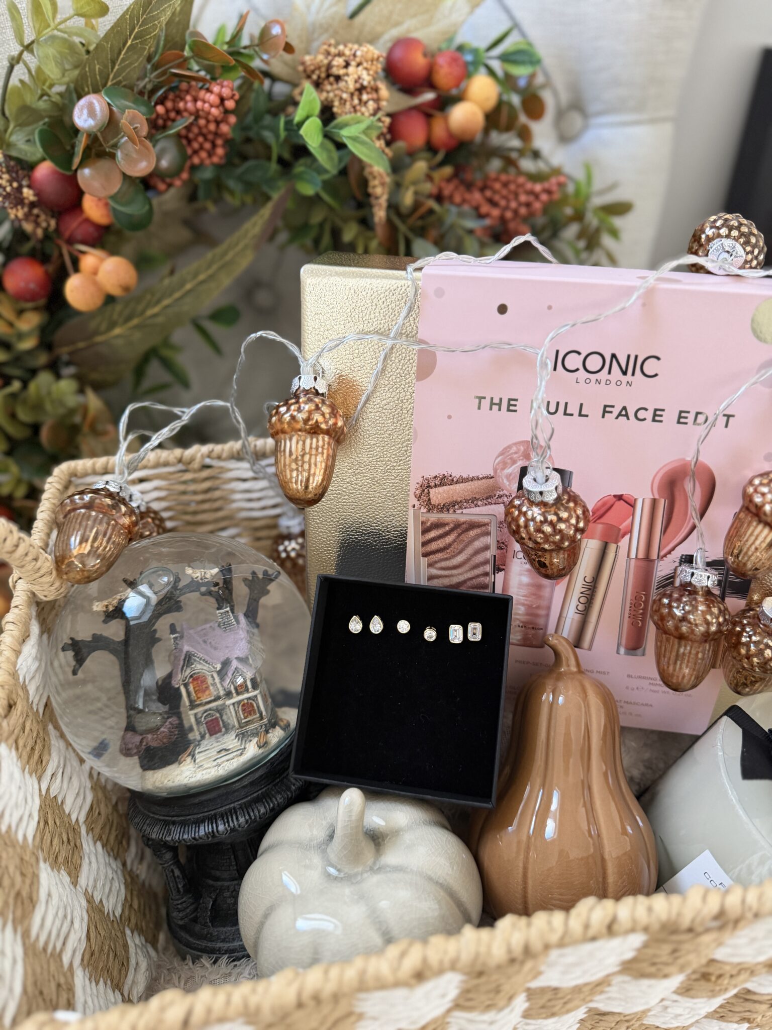 A charming boo basket filled with autumn-themed decor, a snow globe, two ceramic pumpkins, a box of make-up, acorn string lights, stud earrings, and artificial autumn foliage in the background—perfect as a cosy gift or thoughtful autumn present.