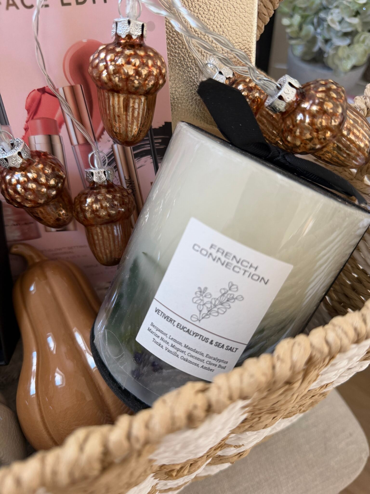 A Boo Basket for her, this charming arrangement contains a French Connection candle, a garland of artificial acorns, a ceramic leaf, and a pink box, with part of a plant visible in the background—an ideal Halloween gift that combines seasonal elegance and thoughtful surprises.