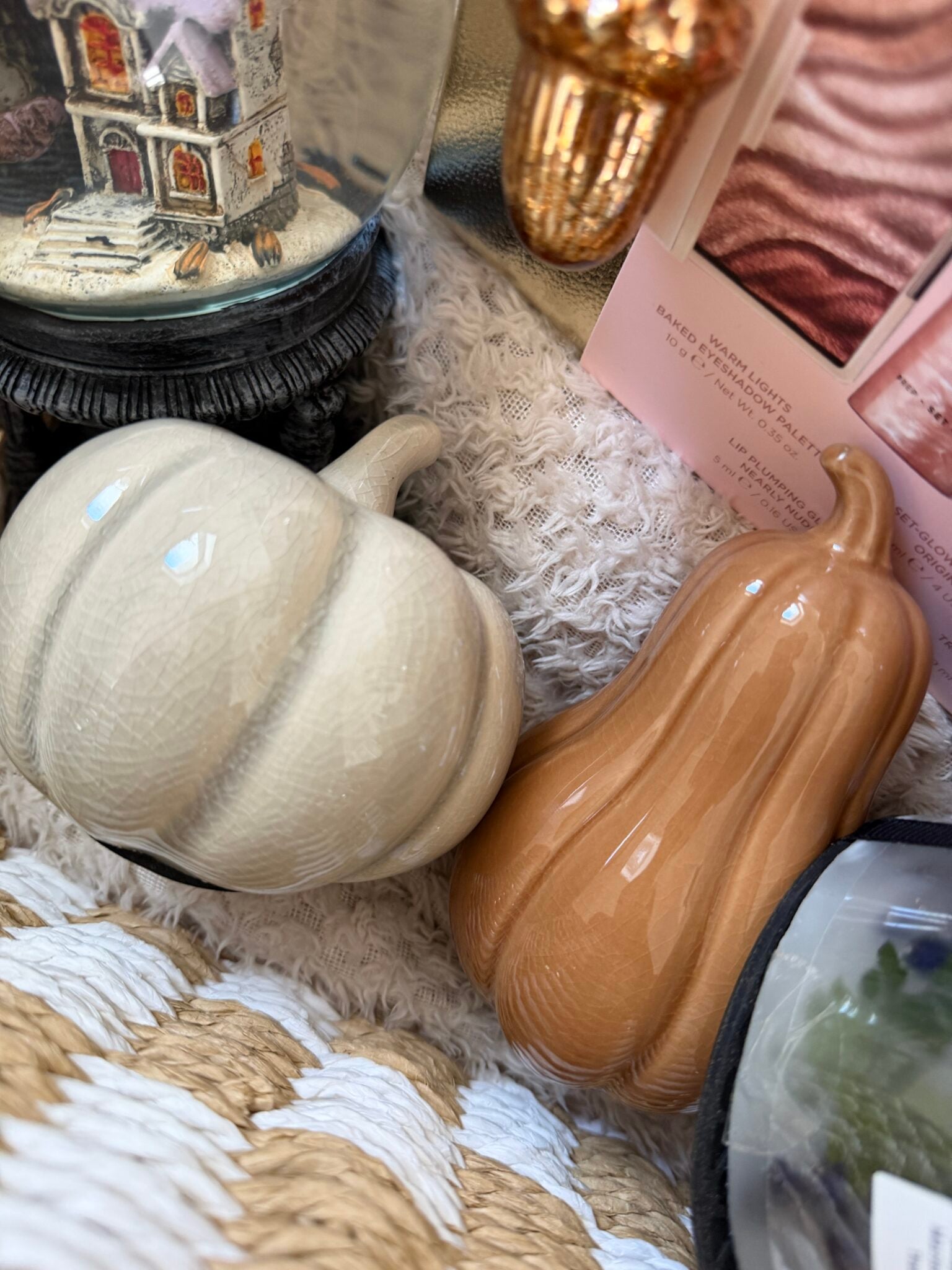 Two ceramic gourds, one white and one light brown, rest on a textured cream blanket next to a few decorative items and pink cards—creating a charming Boo Basket perfect as a Halloween present for her.
