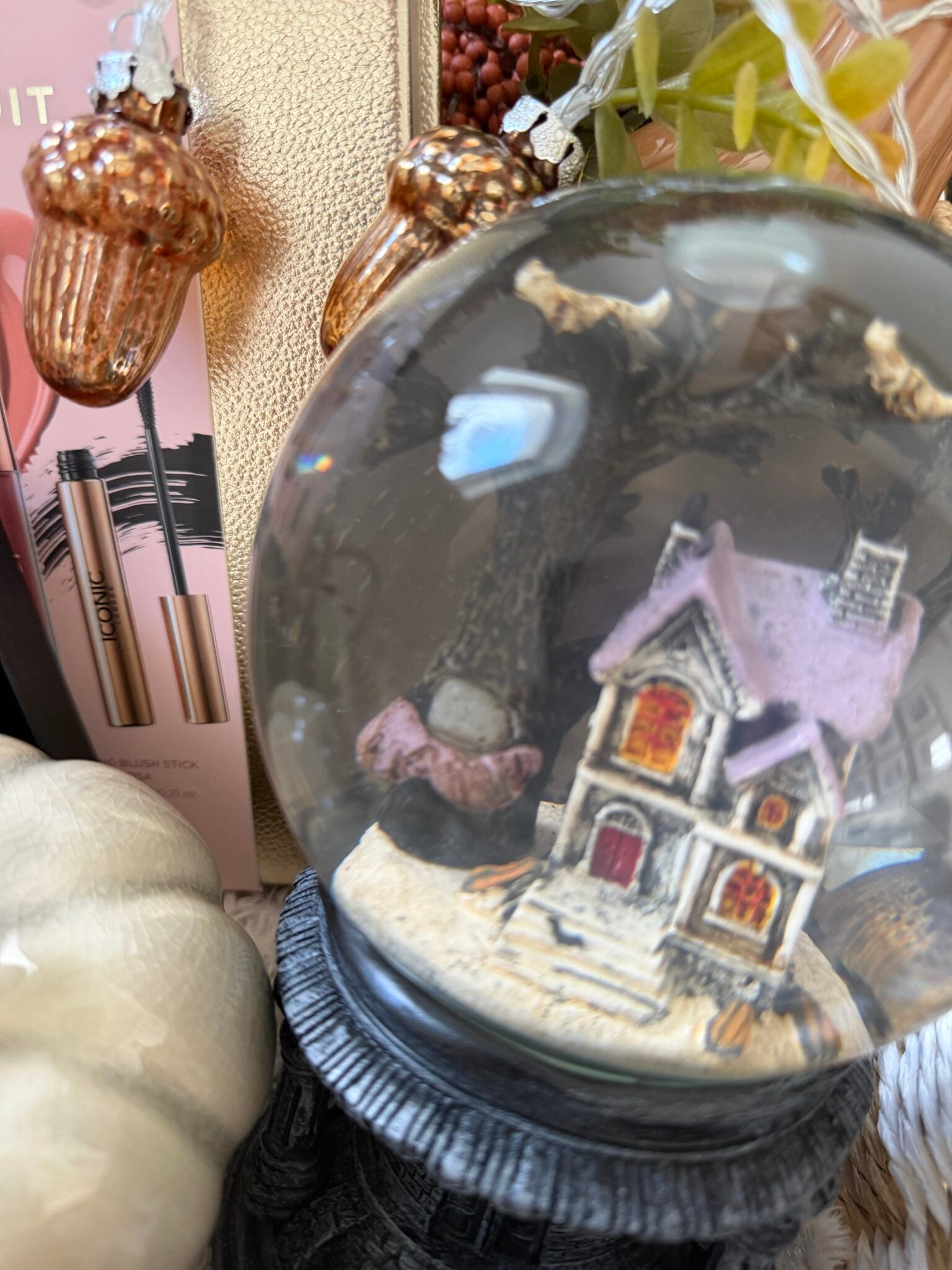 A close-up of a snow globe with a house inside, surrounded by decorative pumpkins, acorn ornaments, and packaged make-up items—perfect for assembling a cosy gift or adding charm to your Halloween gift basket.