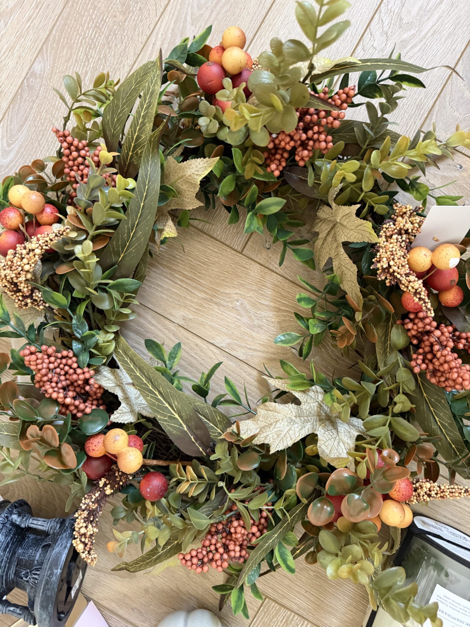 A decorative autumn wreath with green leaves, orange berries, yellow leaves, and small round fruits, placed on a wooden floor—perfect as a cosy gift or autumn present to brighten up any space.