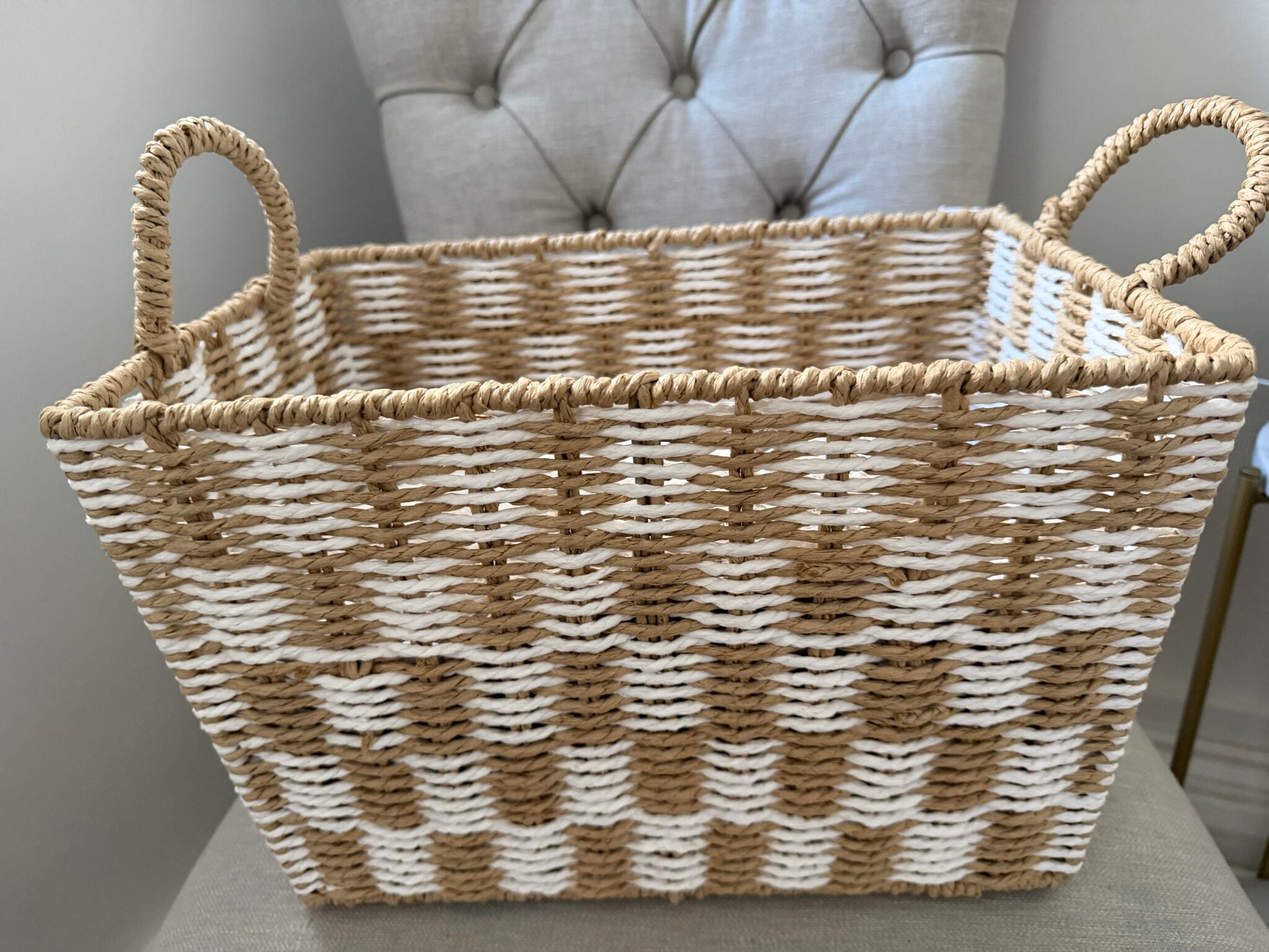 Rectangular woven basket with two handles, featuring a tan and white chequered pattern, placed on a fabric chair with tufted buttons—perfect for creating a cosy autumnal gift or assembling your own Boo Basket.
