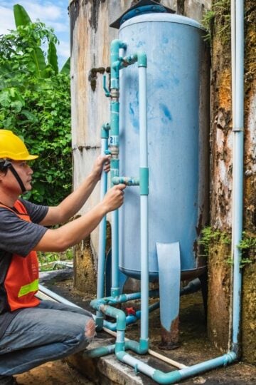 A worker in a yellow helmet and safety vest inspects blue pipes connected to a large vertical metal tank outdoors near a weathered concrete wall, showcasing the importance of regular commercial plumbing maintenance to help prevent unexpected plumbing downtime.