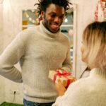 A person in a grey jumper smiles and receives a wrapped present with a red ribbon from another person indoors, capturing the joy of exchanging meaningful gifts for men.