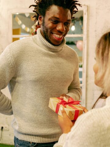 A person in a grey jumper smiles and receives a wrapped present with a red ribbon from another person indoors, capturing the joy of exchanging meaningful gifts for men.