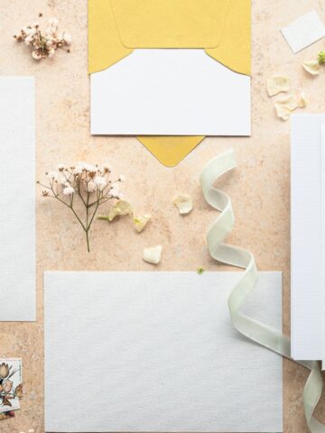 Flat lay of blank white cards, a yellow envelope, dried flowers, a white ribbon, and paper scraps on a beige surface-perfect inspiration for designing family celebrations in 2026 or creating memorable stationery with the help of online invitation platforms.