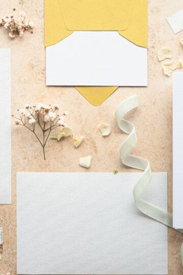 Flat lay of blank white cards, a yellow envelope, dried flowers, a white ribbon, and paper scraps on a beige surface—perfect inspiration for designing family celebrations in 2026 or creating memorable stationery with the help of online invitation platforms.