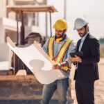 Two men at a construction site review blueprints; one wears a suit and hard hat using a tablet, the other wears safety gear and holds the plans.