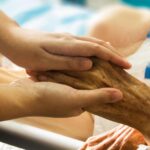A younger person gently holds the hand of an older individual, showing support and care in a setting that appears to be a healthcare or hospital environment-highlighting the importance of palliative care in improving quality of life after a major stroke.