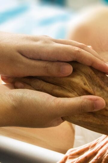 A younger person gently holds the hand of an older individual, showing support and care in a setting that appears to be a healthcare or hospital environment—highlighting the importance of palliative care in improving quality of life after a major stroke.