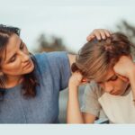 A woman gently comforts a sad child, placing her hand on the child's head while the child looks down with hands on their temples-capturing a tender moment between parents and children as they face the daily battle of navigating school challenges together.