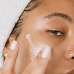 Person applying white cream to their cheek with their fingers, wearing a white towel on their head. The image is a close-up focusing on skin and facial care, highlighting the importance of keeping flaky skin hydrated.