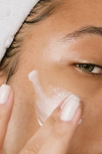 Person applying white cream to their cheek with their fingers, wearing a white towel on their head. The image is a close-up focusing on skin and facial care, highlighting the importance of keeping flaky skin hydrated.