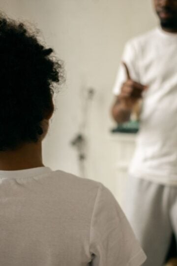 A child faces an adult who is pointing and appears to be speaking. Both are wearing white shirts and standing indoors in a softly lit room.