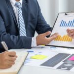 Two people in business attire discuss graphs and charts at a table, with one person holding a clipboard displaying bar and line graphs to better understand customer behaviour in their business.